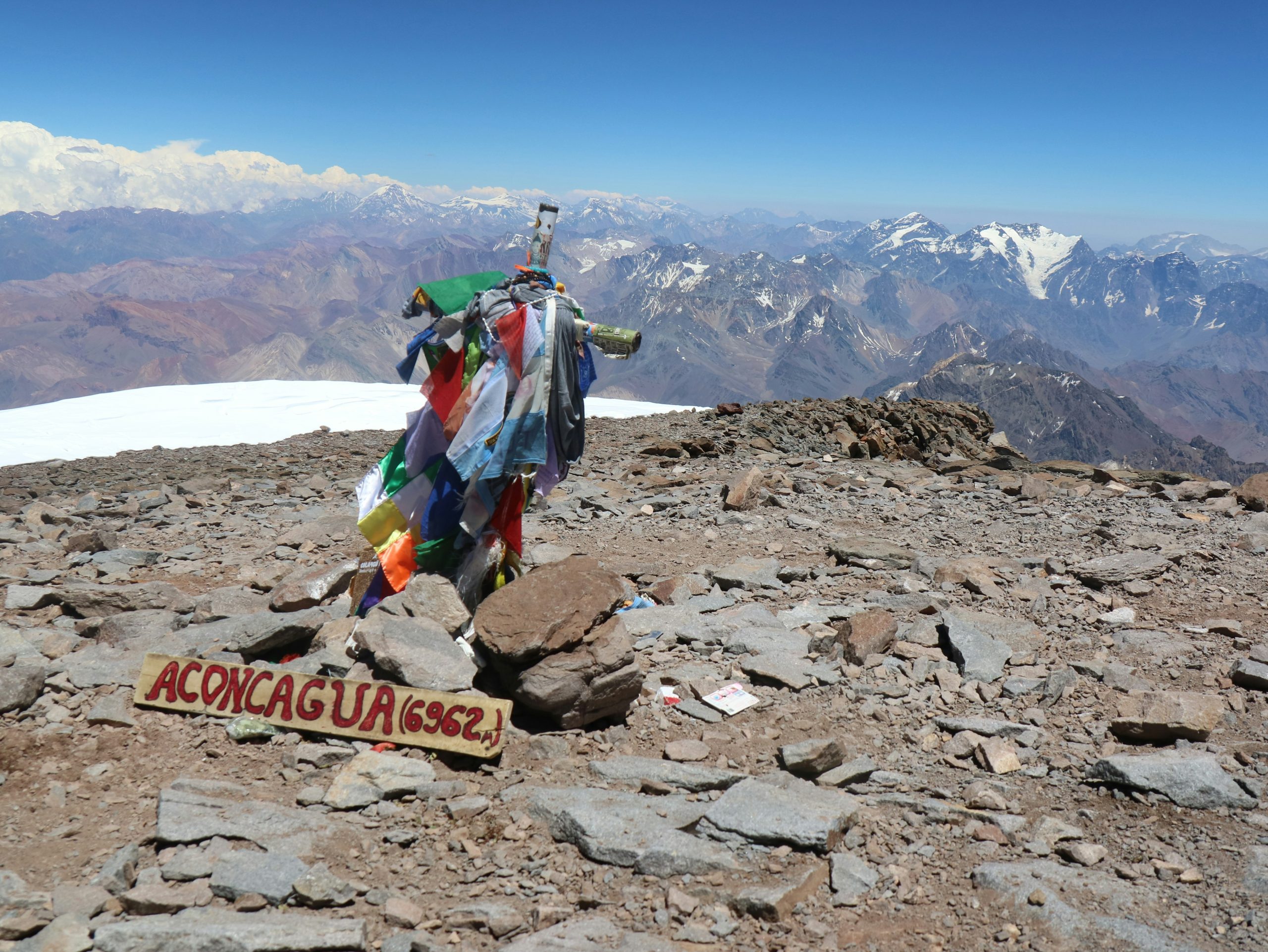 Aconcagua summit sign at 6,962 m, with prayer flags and a panoramic view of the Andes